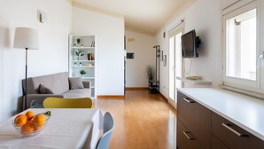 A modern living room with a sofa, a dining table with a bowl of fruit, a white cabinet with a TV, and a bookshelf. - Attico Eleonora – Terrazza & Parcheggio in Centro (Oristano)