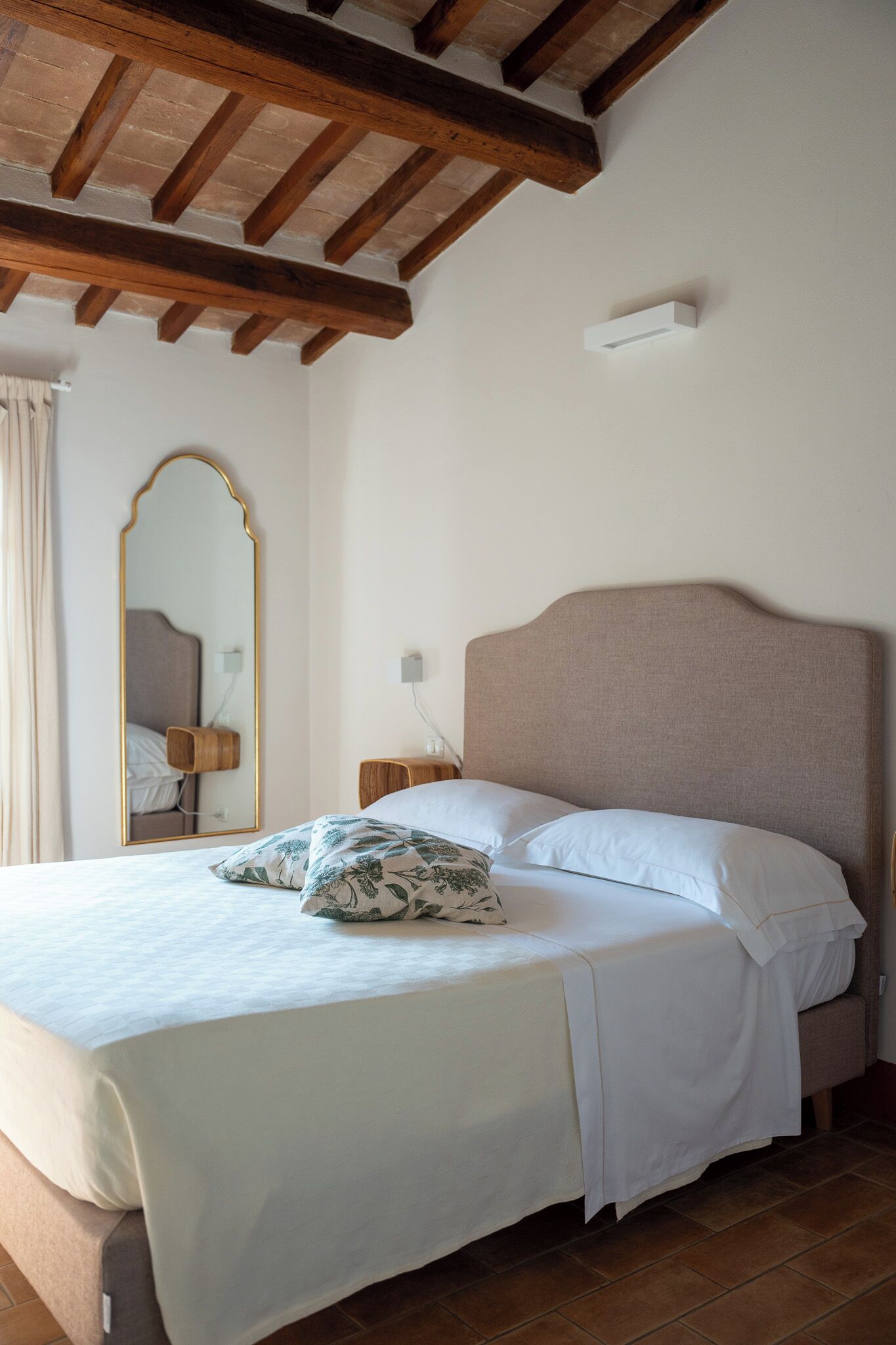 A neatly made bed with white linens and a floral pillow, a wooden headboard, a large mirror on the wall, and a small bedside table with a lamp.