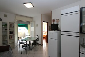 A compact kitchen with a refrigerator, a dining table with chairs, and a small TV on top of the fridge. - Comfy Flat by the Beach (San Michele al Tagliamento)