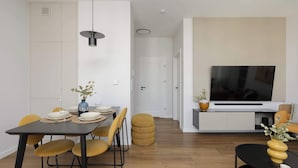 Un salon moderne avec un coin repas, comprenant une table à manger noire, des chaises jaunes, une table basse noire et un téléviseur fixé sur un meuble blanc.