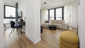 Un salon moderne avec un canapé, une table basse et un fauteuil. On y trouve un bureau avec une chaise et un téléviseur. La pièce est dotée de grandes fenêtres avec des rideaux.