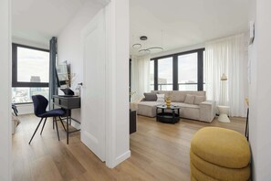 Ein modernes Wohnzimmer mit einer Couch, einem Couchtisch und einem Sessel. Ein Schreibtisch mit Stuhl und ein Fernseher stehen ebenfalls im Raum. Große Fenster mit Vorhängen sorgen für viel Licht.