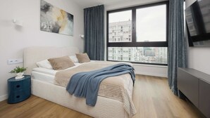 Une chambre moderne avec un grand lit, une vue sur la ville, un téléviseur  à écran plat et une table de chevet agrémentée d’une plante.