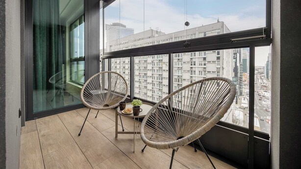 Un balcon avec deux fauteuils en osier, une petite table ornée d’une plante et d’une tasse, et une vue sur le paysage urbain à travers de grandes fenêtres.