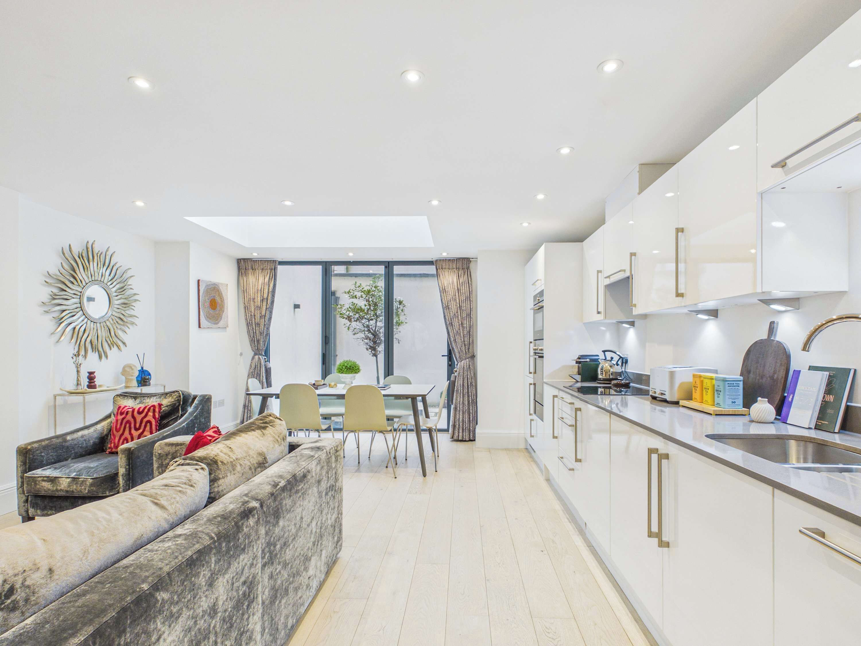 A modern kitchen with white cabinets, a dining area with a table and chairs, and a living area with a sofa and a decorative mirror.