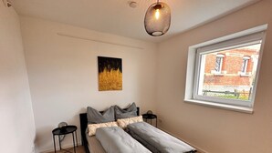 A modern bedroom with a bed, bedside tables, a hanging light fixture, a window with a view of a brick building, and a wall-mounted artwork.