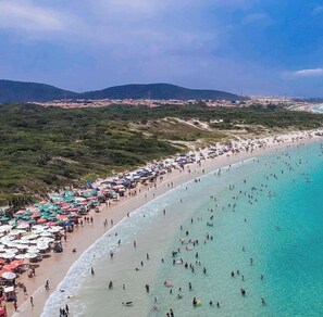 A beach with people swimming and sunbathing, surrounded by greenery and hills. - Comfortable house—pool, grill, Wi-Fi, air conditioning, linens (Cabo Frio)