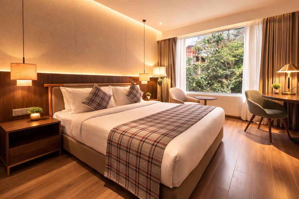 A hotel room with a large bed, bedside lamps, a small table, a chair, and a window with curtains.
