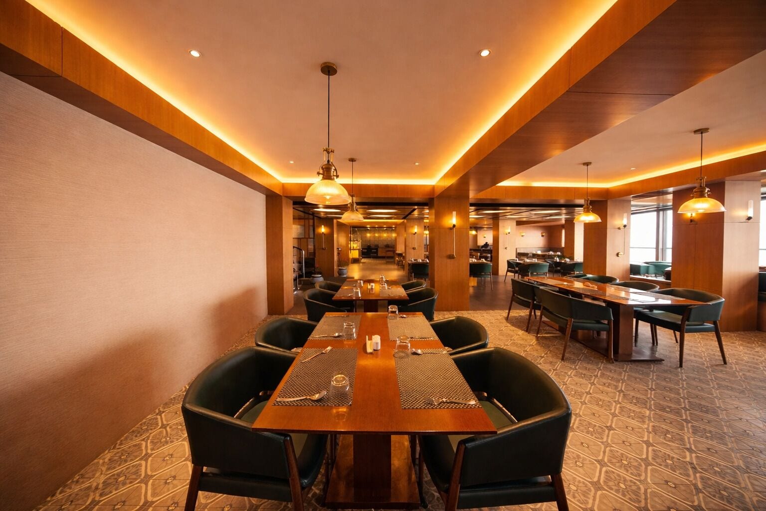 Uma sala de jantar de hotel com mesas compridas, cadeiras e luminárias pendentes.