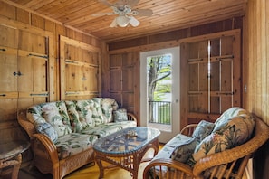 Un intérieur en bois comprenant un canapé, des fauteuils en osier, une table avec un plateau en verre et un ventilateur de plafond.