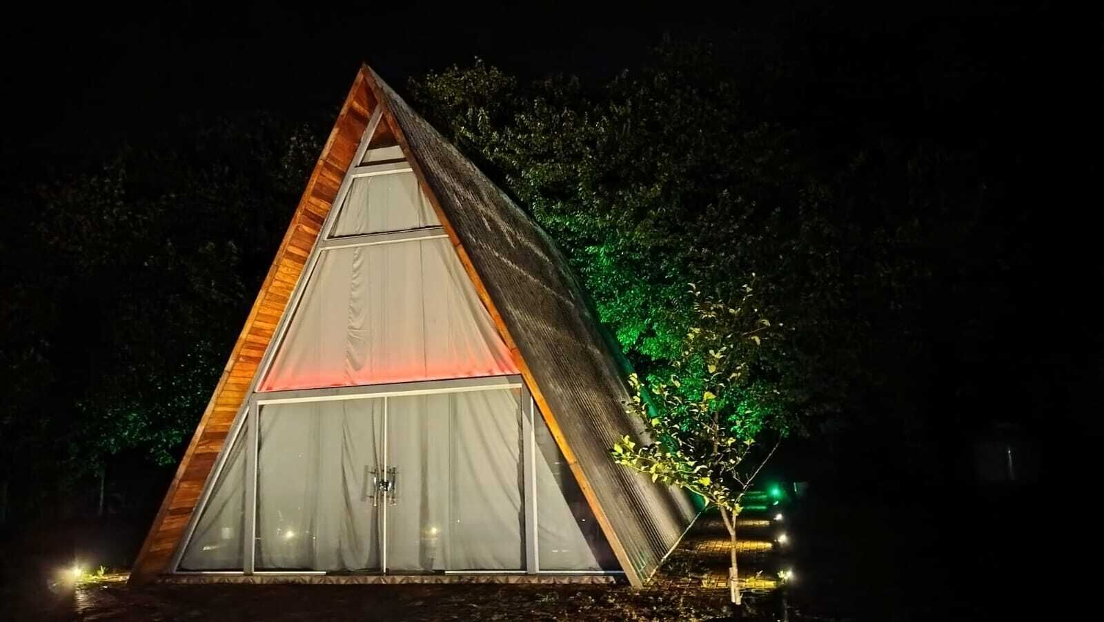 Una struttura a forma di tenda triangolare, con un'apertura centrale e una più piccola alla base, circondata da alberi e illuminata da luci artificiali.
