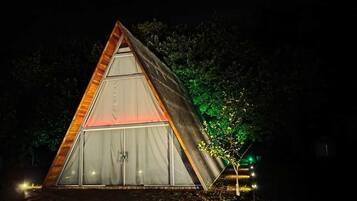 Una struttura a forma di tenda triangolare, con un'apertura centrale e una più piccola alla base, circondata da alberi e illuminata da luci artificiali.