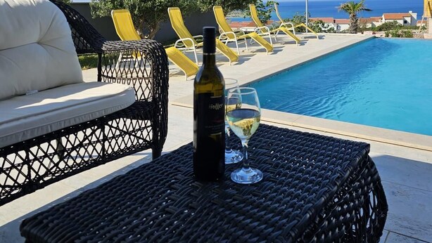 Uma área à beira da piscina com espreguiçadeiras, uma mesa de vime com uma garrafa e um copo, e vista para o mar.