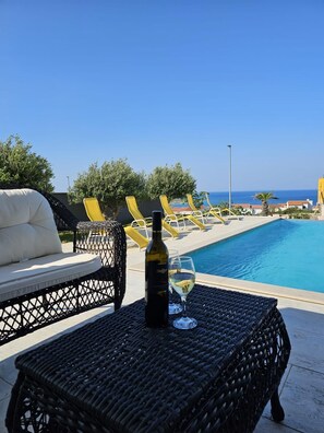 Ein Poolbereich mit Liegestühlen, einem Korbstuhl-Tisch, einer Flasche und einem Glas sowie Blick auf das Meer.