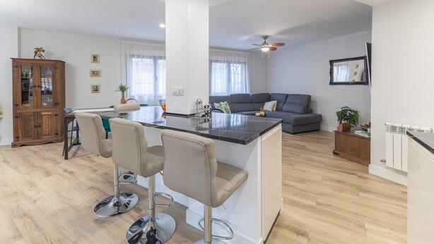 Eine moderne Küche mit einer zentralen Kochinsel, Barhocker, einem Esstisch und einem grauen Sofa im Wohnbereich.