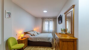 Ein Schlafzimmer mit Bett, Nachttisch, Lampe, Sessel, Kommode, Spiegel, Fernseher und Vorhängen.