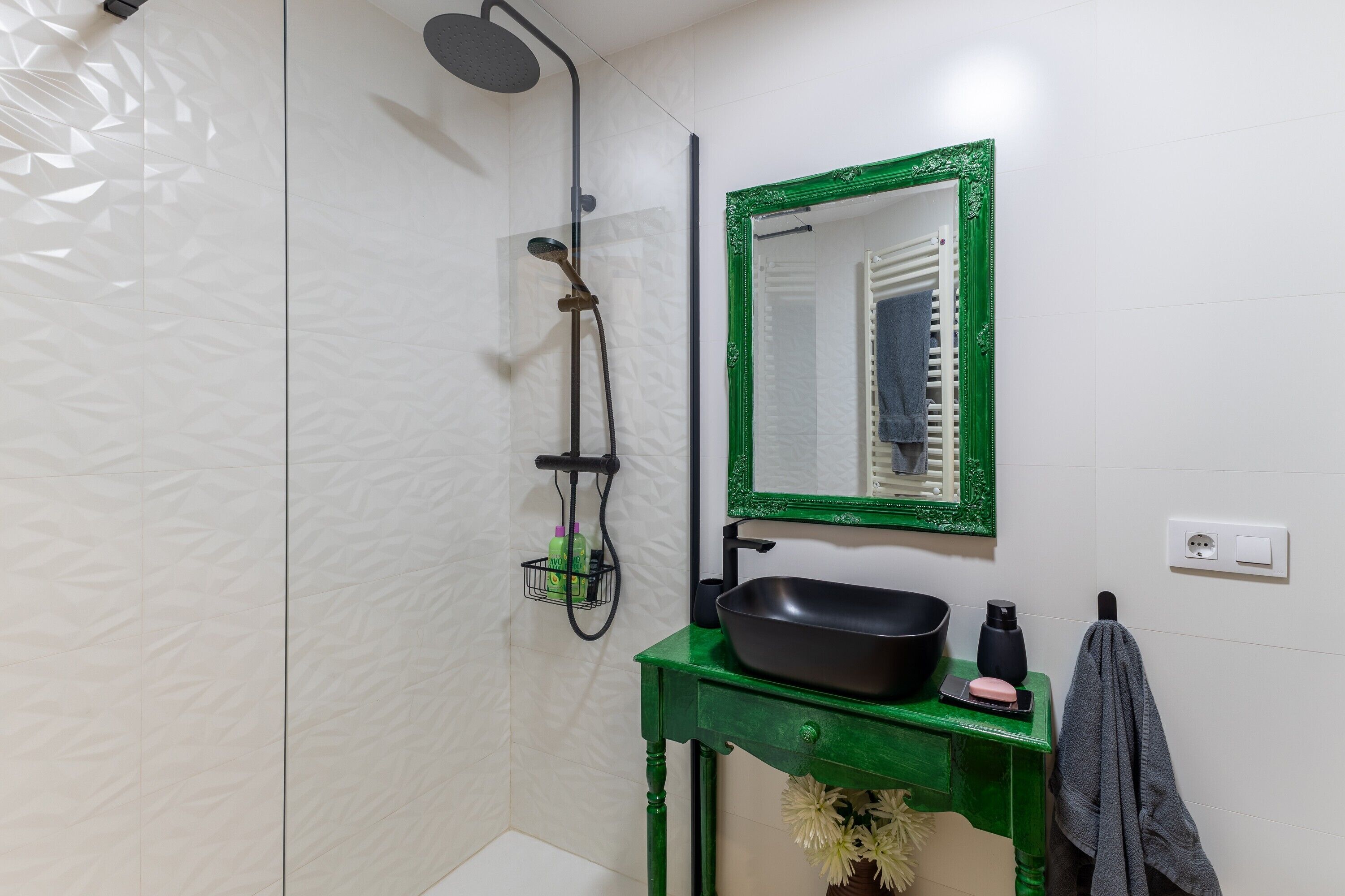 Un baño con ducha, un mueble de tocador verde, un lavabo negro, un espejo con marco verde y un perchero.