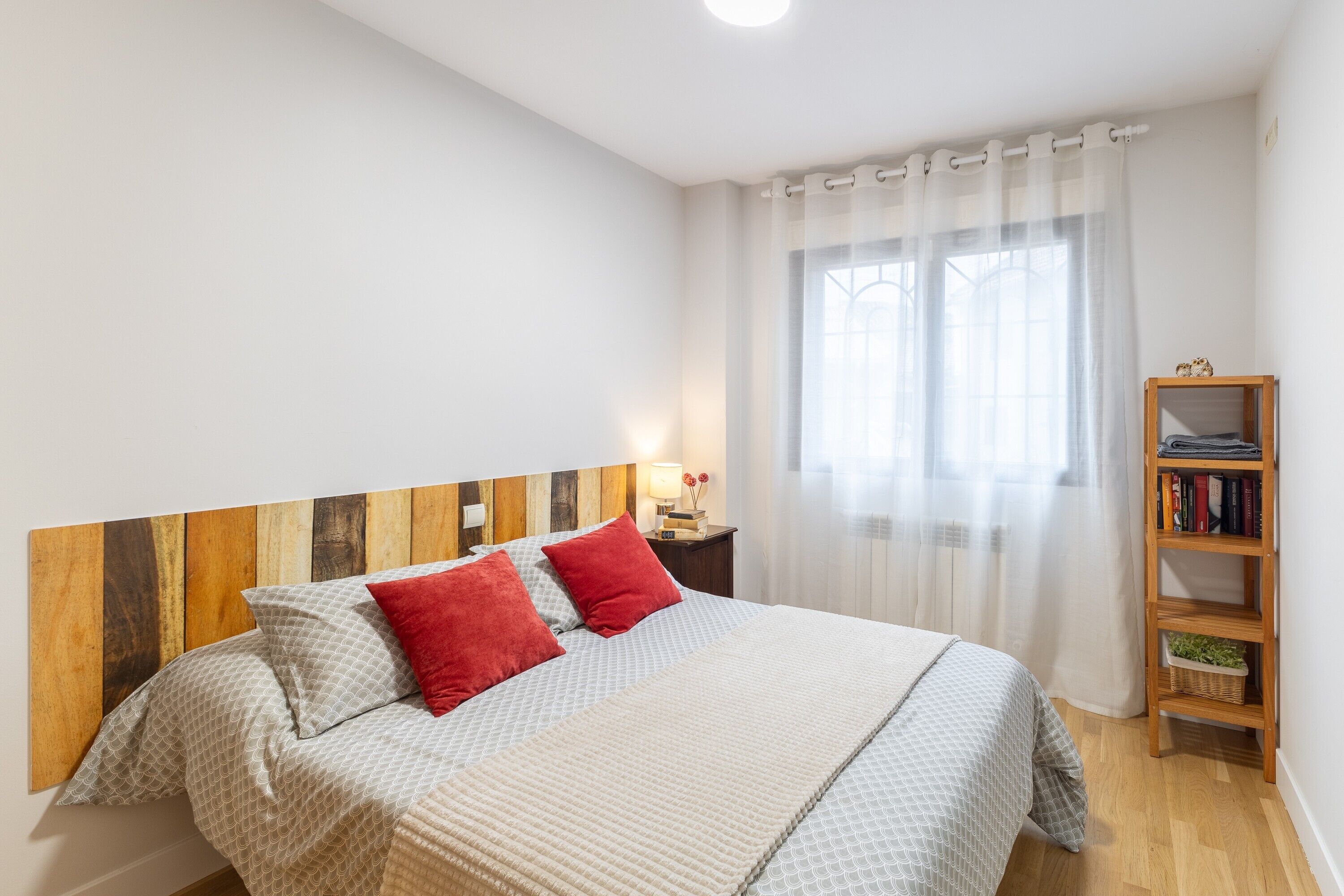 Ein Schlafzimmer mit hölzernem Kopfteil, einem Bett mit gemusterten Bettbezügen, roten Kissen, einem Nachttisch mit Lampe, einem Fenster mit leichtem Vorhang und einem Bücherregal aus Holz.
