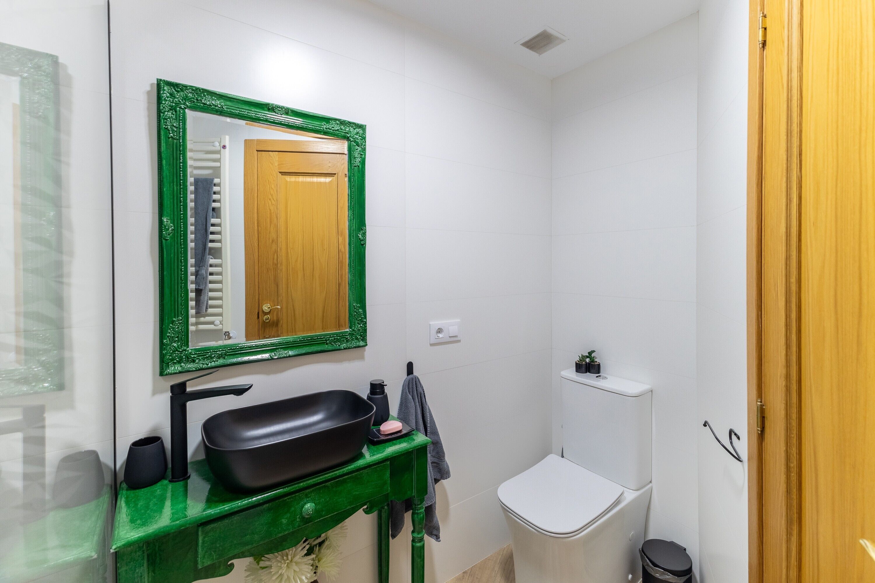 Een badkamer met een groen badkamerkastje, een zwarte wastafel, een spiegel met groene lijst, een wit toilet en een klein plankje met toiletartikelen.