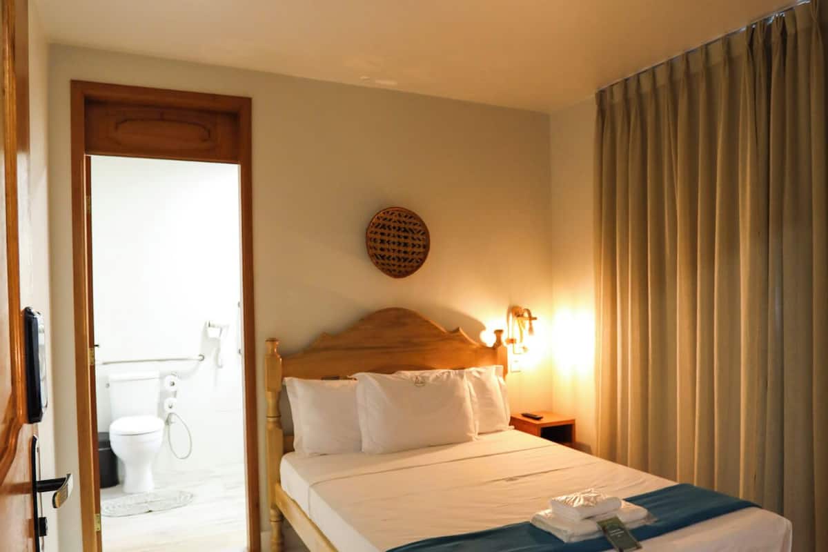 A bedroom with a wooden bed, a bedside table, a wall-mounted light, and a view of the bathroom.