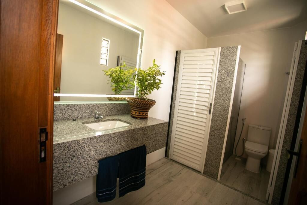 Un baño con encimera de granito, un espejo, un lavabo, una planta en maceta, un perchero para toallas y un inodoro.