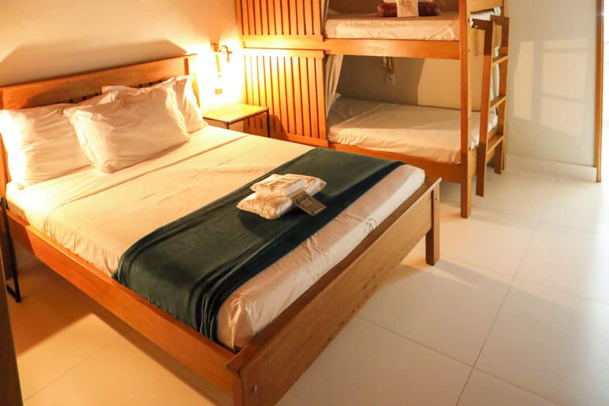 A room with a wooden bunk bed, a single bed, a bedside table with a lamp, and a wall-mounted fan.