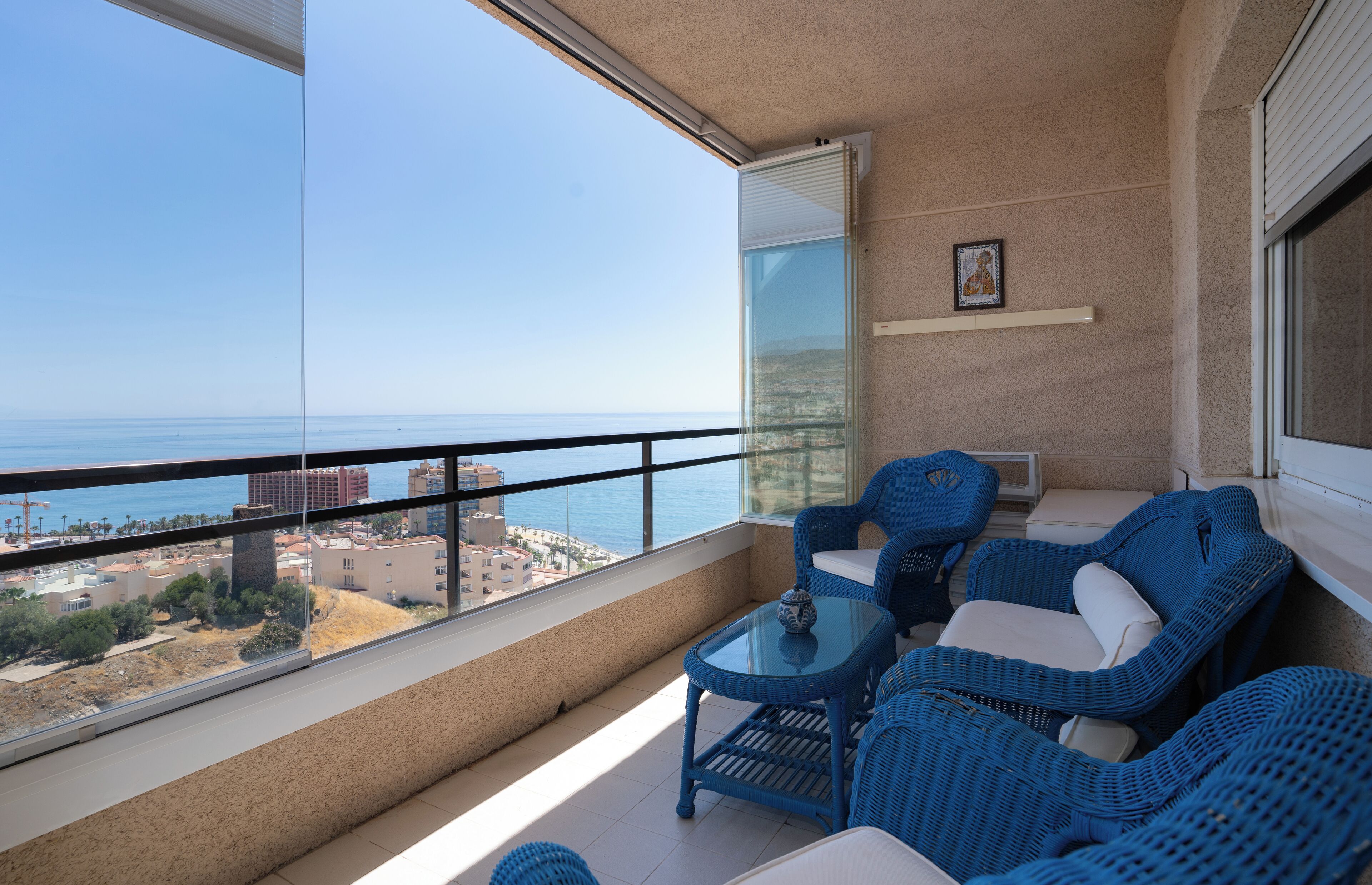 Ein Balkon mit Korbmöbeln, einem Glastisch und Blick auf das Meer und die Bebauung.