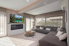 A modern living room with a flat-screen TV mounted on the wall, a grey sofa, a coffee table with a tray of muffins, and a large window with sheer curtains. - Villa Centinera With Private Pool (Medulin)