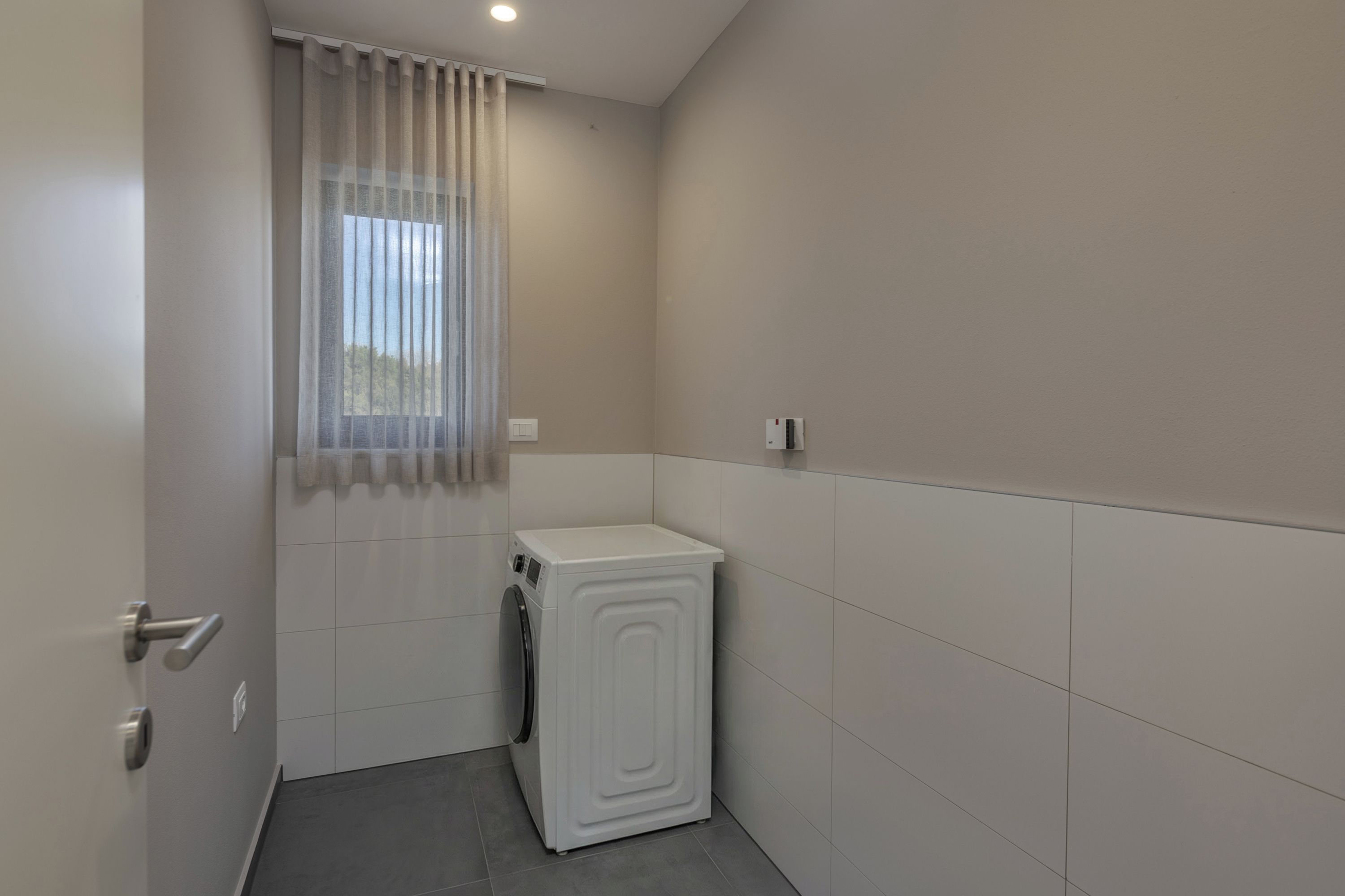 A compact laundry room with a washing machine, a door with a handle, and a window with sheer curtains.