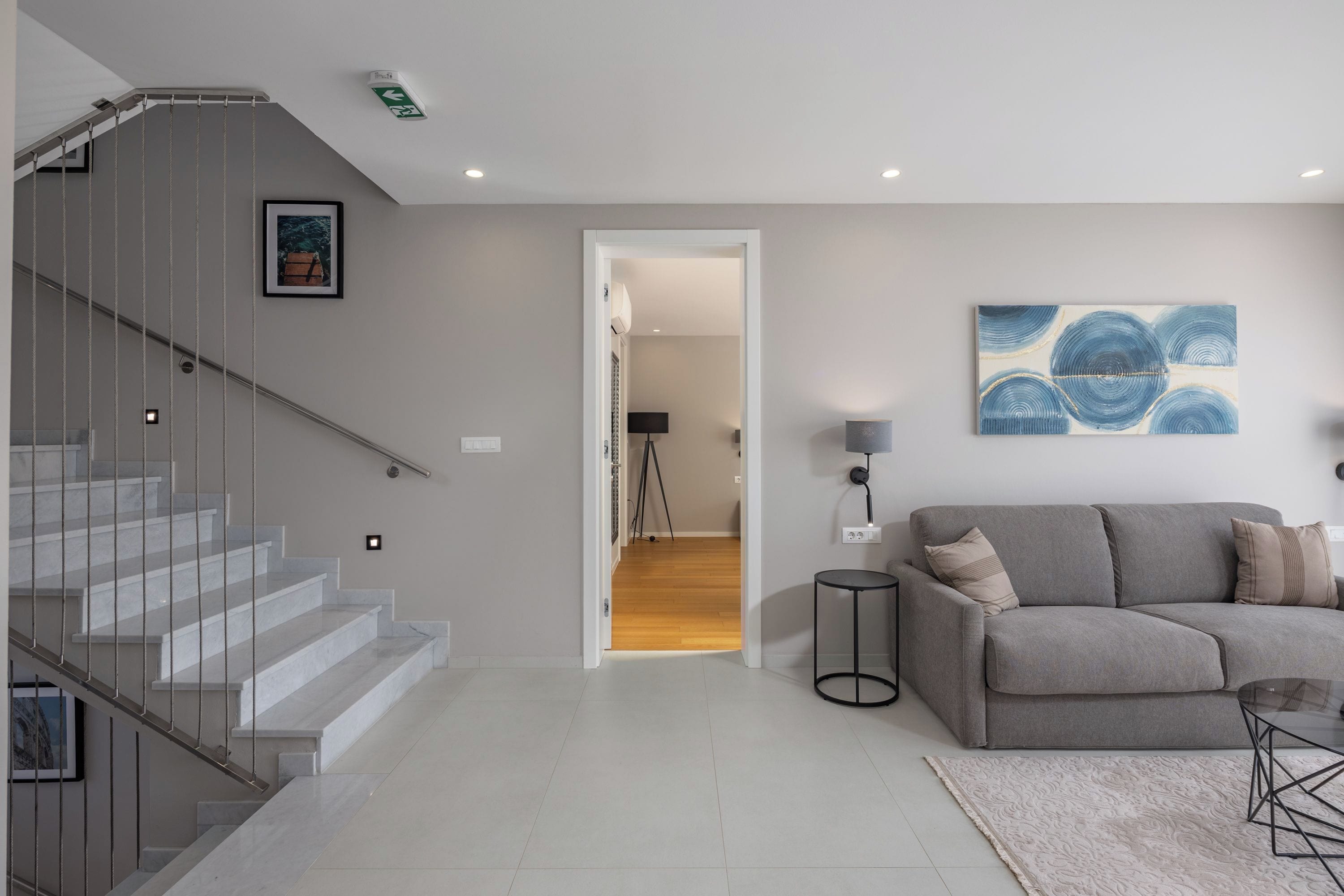 A modern living room with a staircase, a grey sofa, a coffee table, and a painting on the wall.
