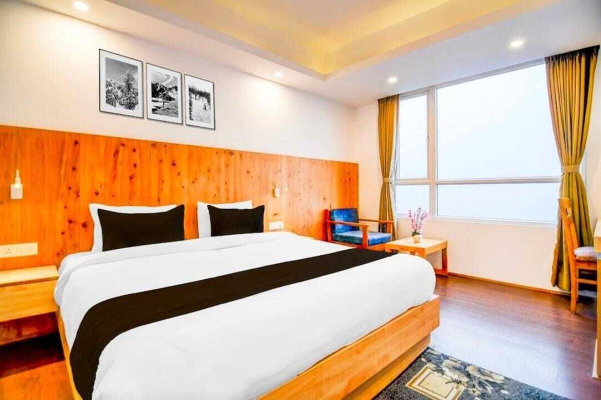 A hotel room with a large bed, wooden headboard, a small table with a chair, and a window with curtains.