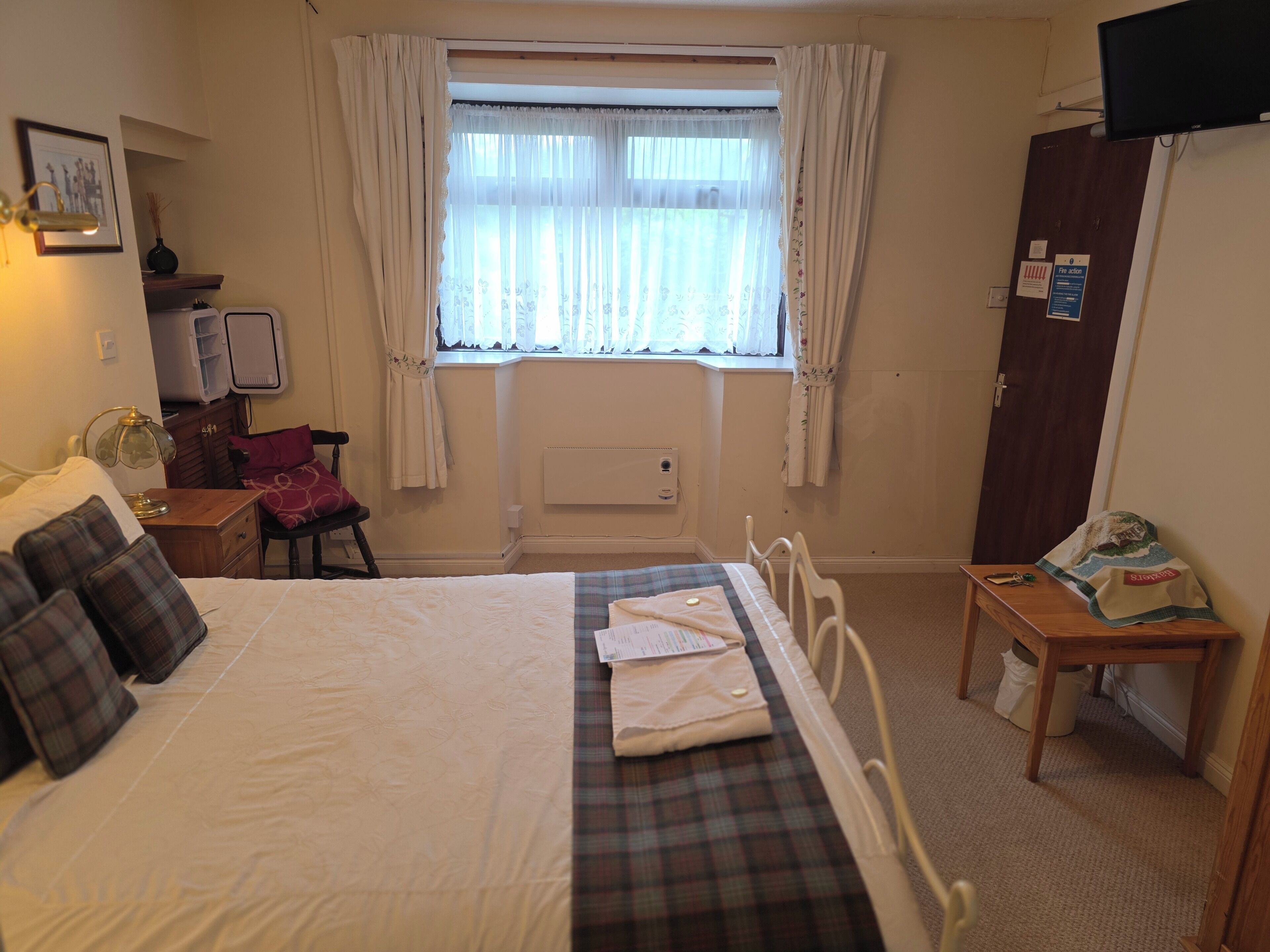A bedroom with a bed, a desk, a chair, a window with curtains, a radiator, and a television.