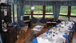 A dining area with a long table set for a meal, a sofa with cushions, a television, and a bar area with bottles. - Achnabobane Farmhouse (Spean Bridge)