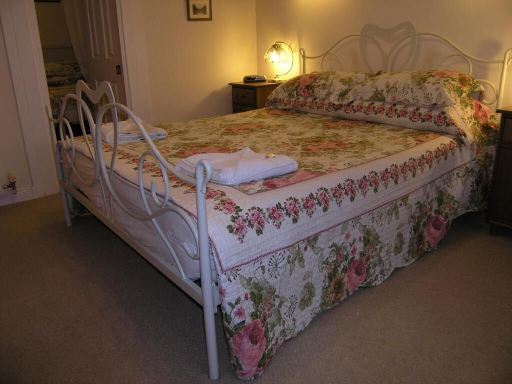 Una cama individual con colcha estampada y estructura metálica blanca. Hay una mesita de noche con una lámpara y una toalla doblada sobre la cama.