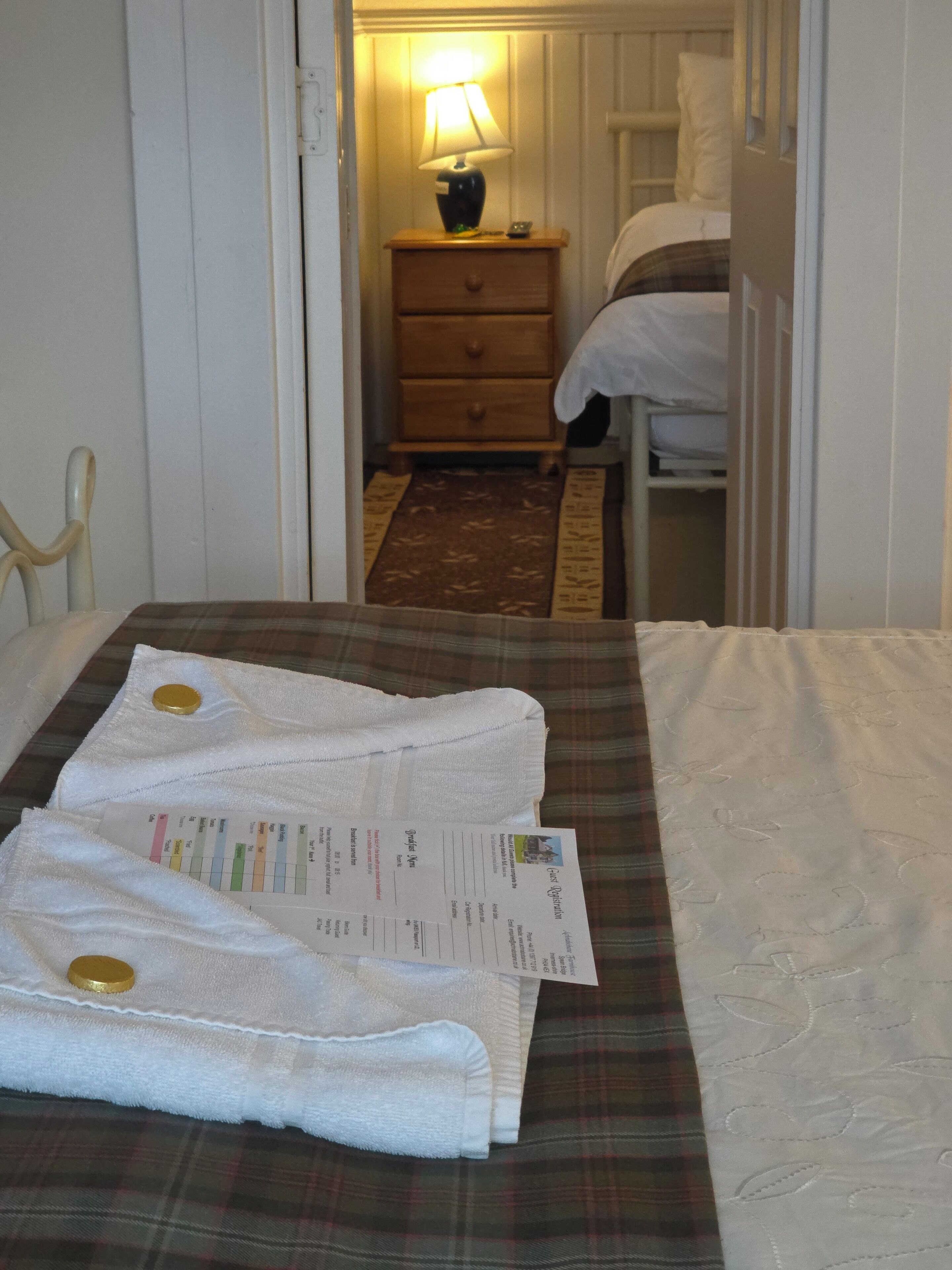 A hotel room with a bed, a nightstand with a lamp, a wooden dresser, and a check-in form on the bed.