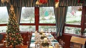 A dining area with a long table set for a meal, decorated with a floral table runner and candles. A Christmas tree is visible in the background, and the room has large windows with curtains. - Achnabobane Farmhouse (Spean Bridge)