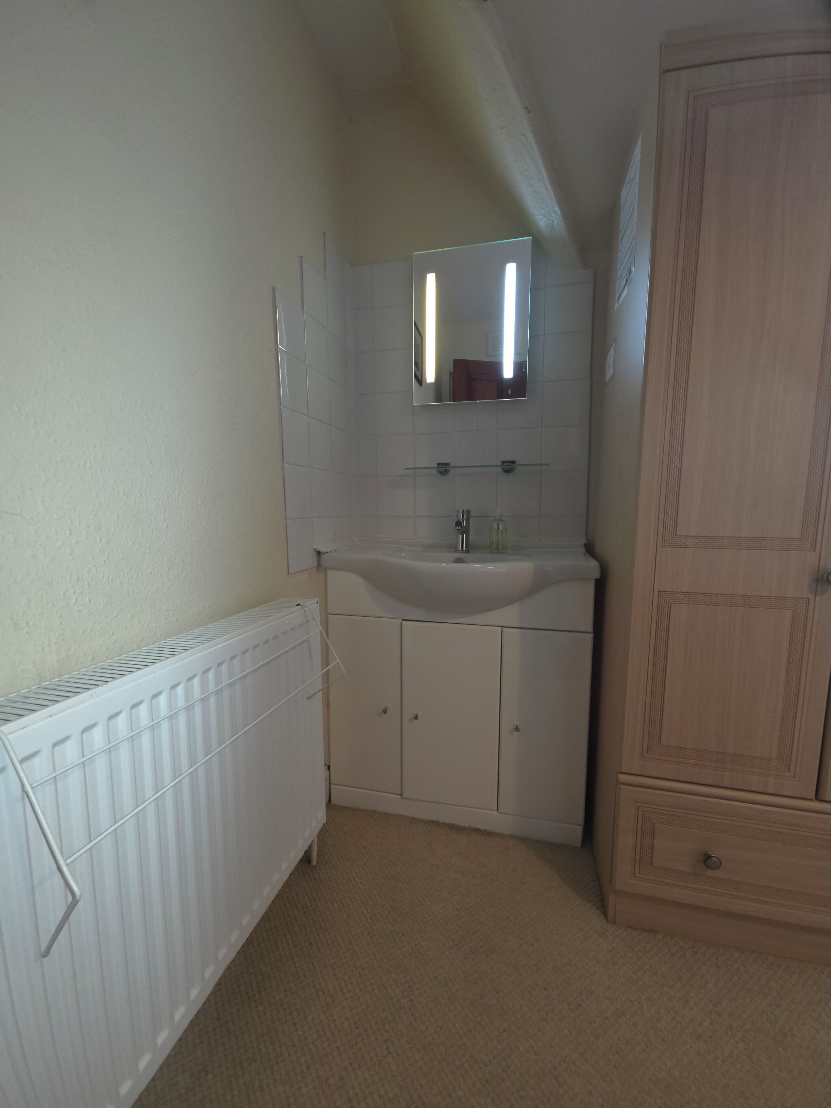 A bathroom with a white radiator, a sink, and a mirror above it. There is a wooden wardrobe to the right.