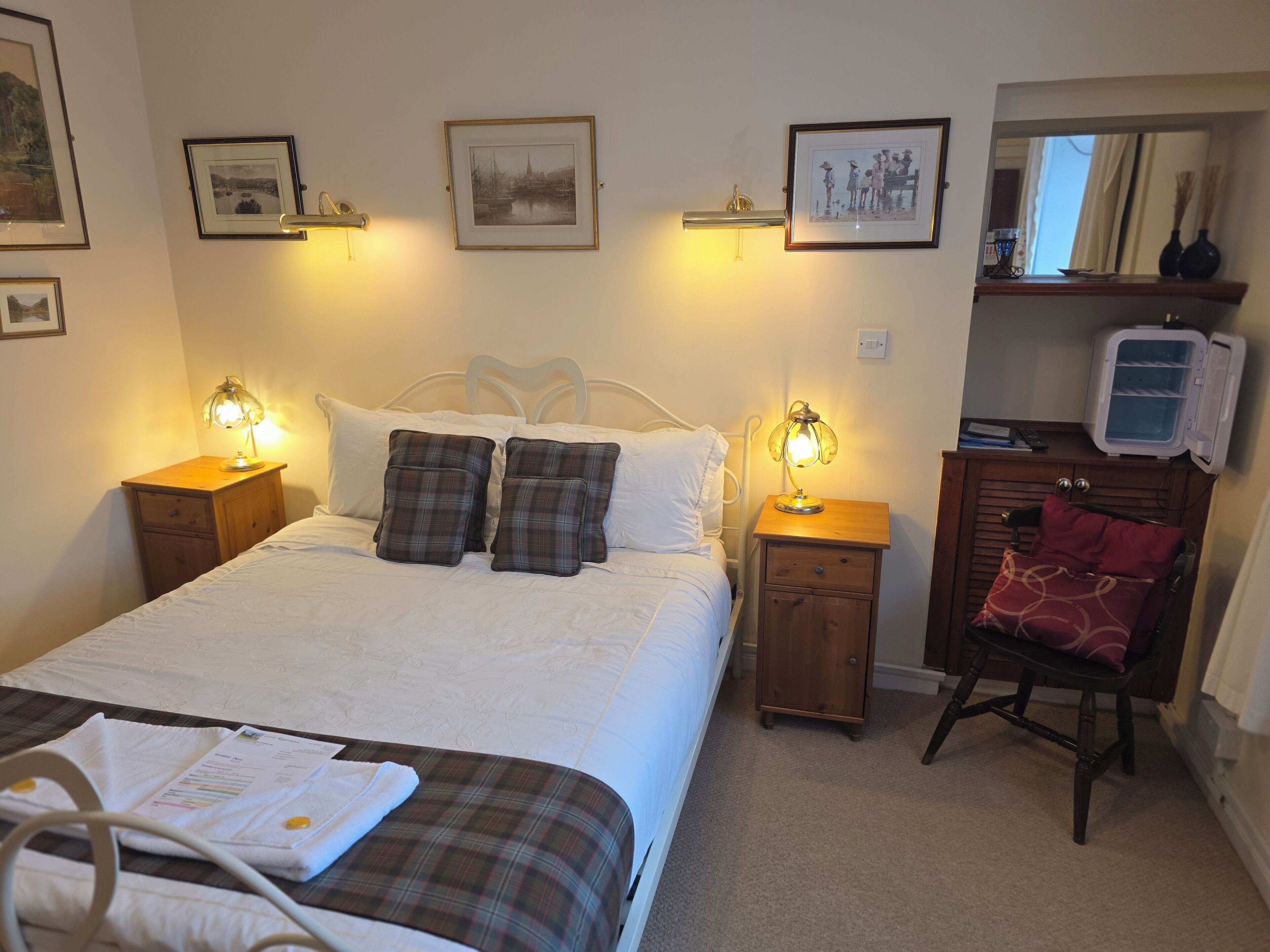 A neatly arranged bedroom with a bed, bedside lamps, a nightstand, a chair, a dresser, and framed pictures on the wall.