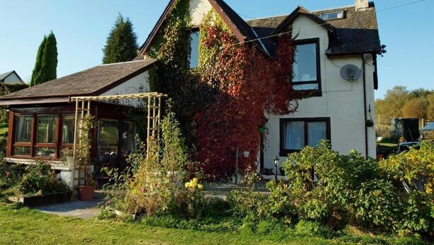 A two-story house with a sloped roof, ivy-covered walls, and a visible satellite dish. - Achnabobane Farmhouse (Spean Bridge)