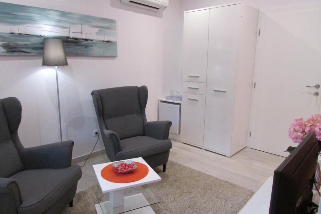 A modern room with two grey armchairs, a white table with a red coaster, a painting on the wall, a wardrobe, and a small side table with a water bottle.