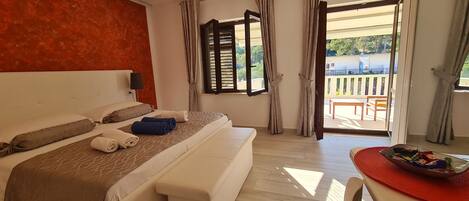 Habitación de hotel con una cama grande, un banco, una mesa con un plato de comida y un balcón con vistas.