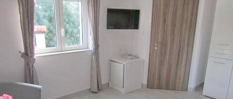 A room with a window, a television, a small white cabinet, and a wardrobe.