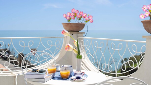 Une table avec un livre, des lunettes et des boissons, un vase rempli de fleurs et une vue sur la mer.