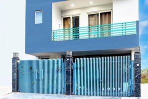 Um edifício moderno de dois andares, com fachada azul, entrada protegida por grades e varanda com guarda-corpo azul.