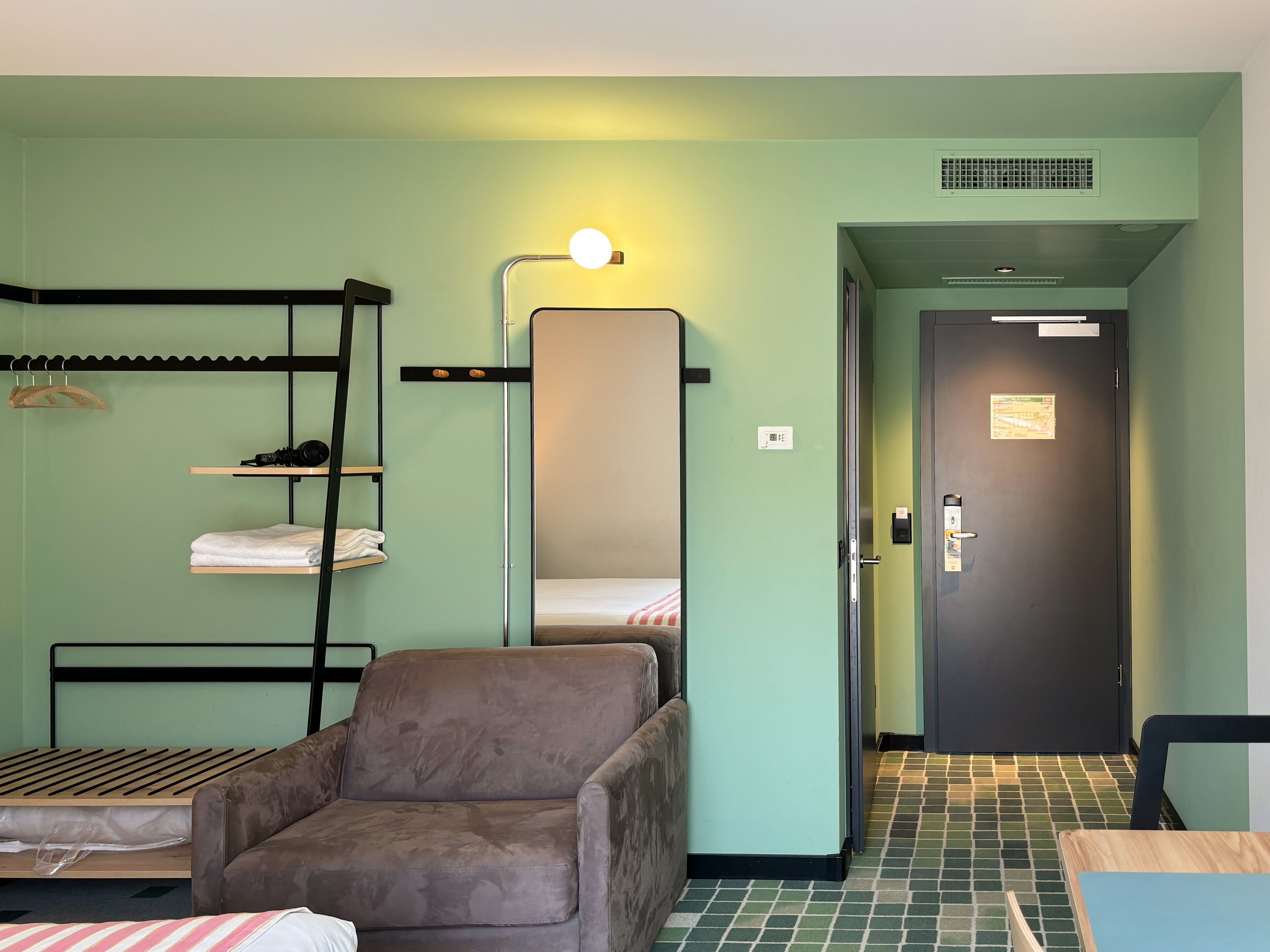 A compact hotel room with bunk beds, a single chair, a small table, and a door leading to another room.