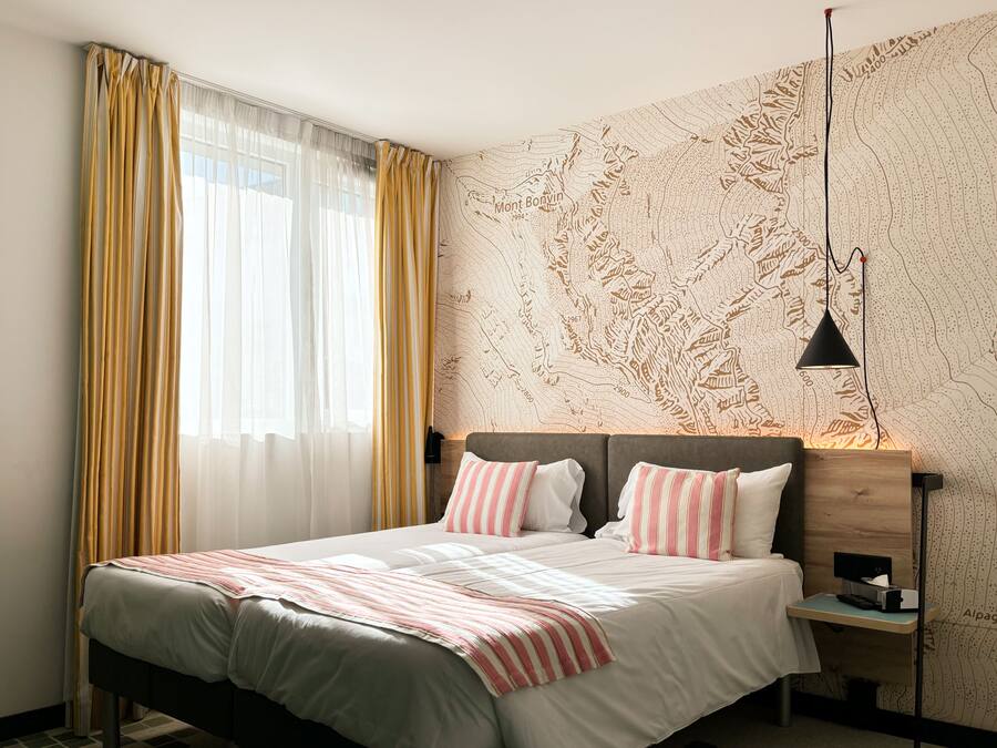 A double bed with striped bedding, a bedside table with a lamp, and a wall with a topographic map design.