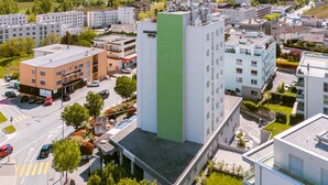 Um edifício moderno com fachada verde, rodeado por outros edifícios e uma paisagem montanhosa ao fundo.