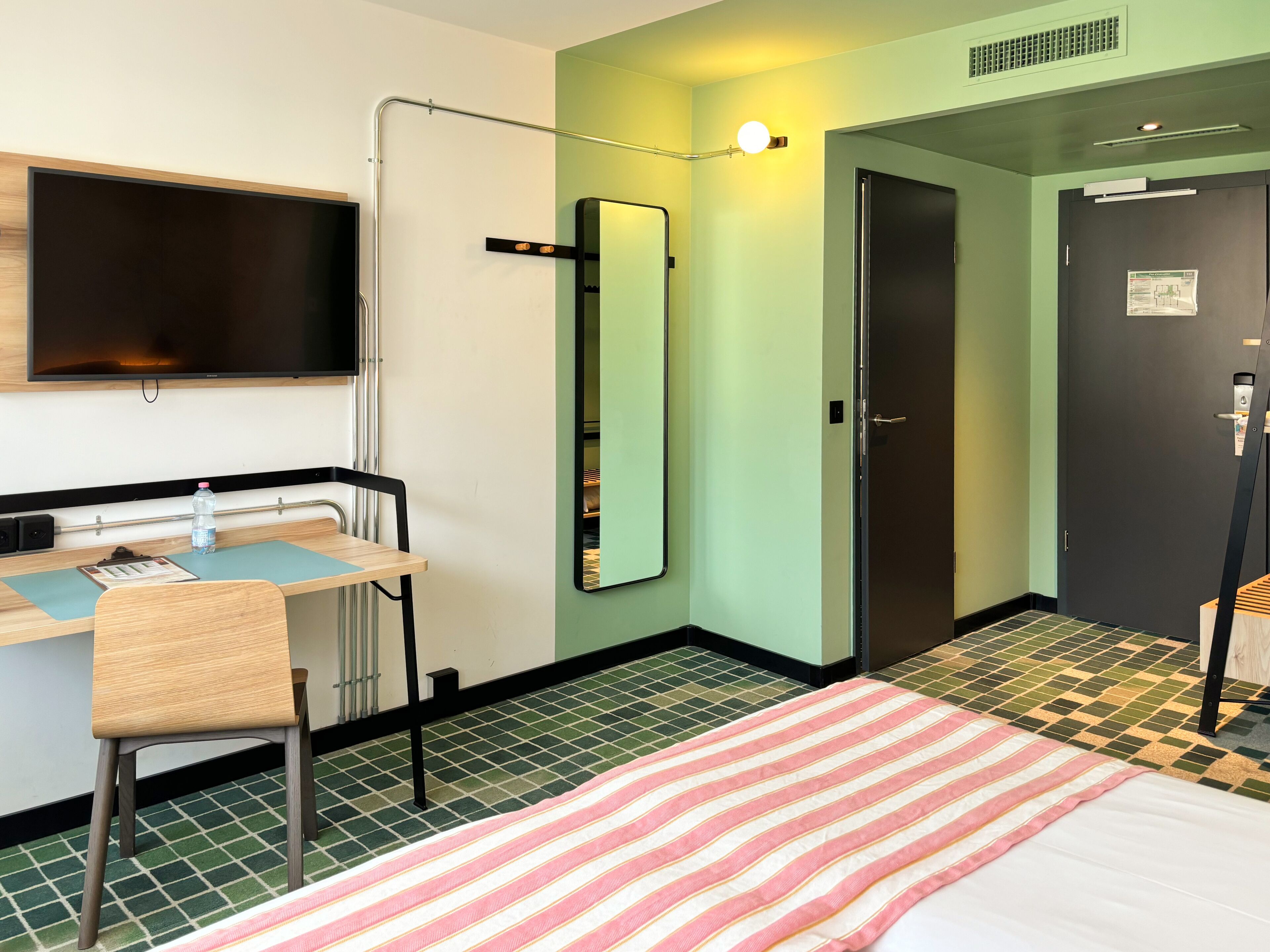 Quarto de hotel com cama, escrivaninha, cadeira, espelho, televisão e porta.