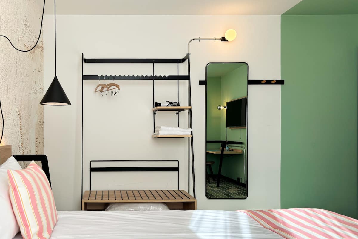 Habitación de hotel compacta con una cama litera, un escritorio pequeño, un espejo y estante en la pared.