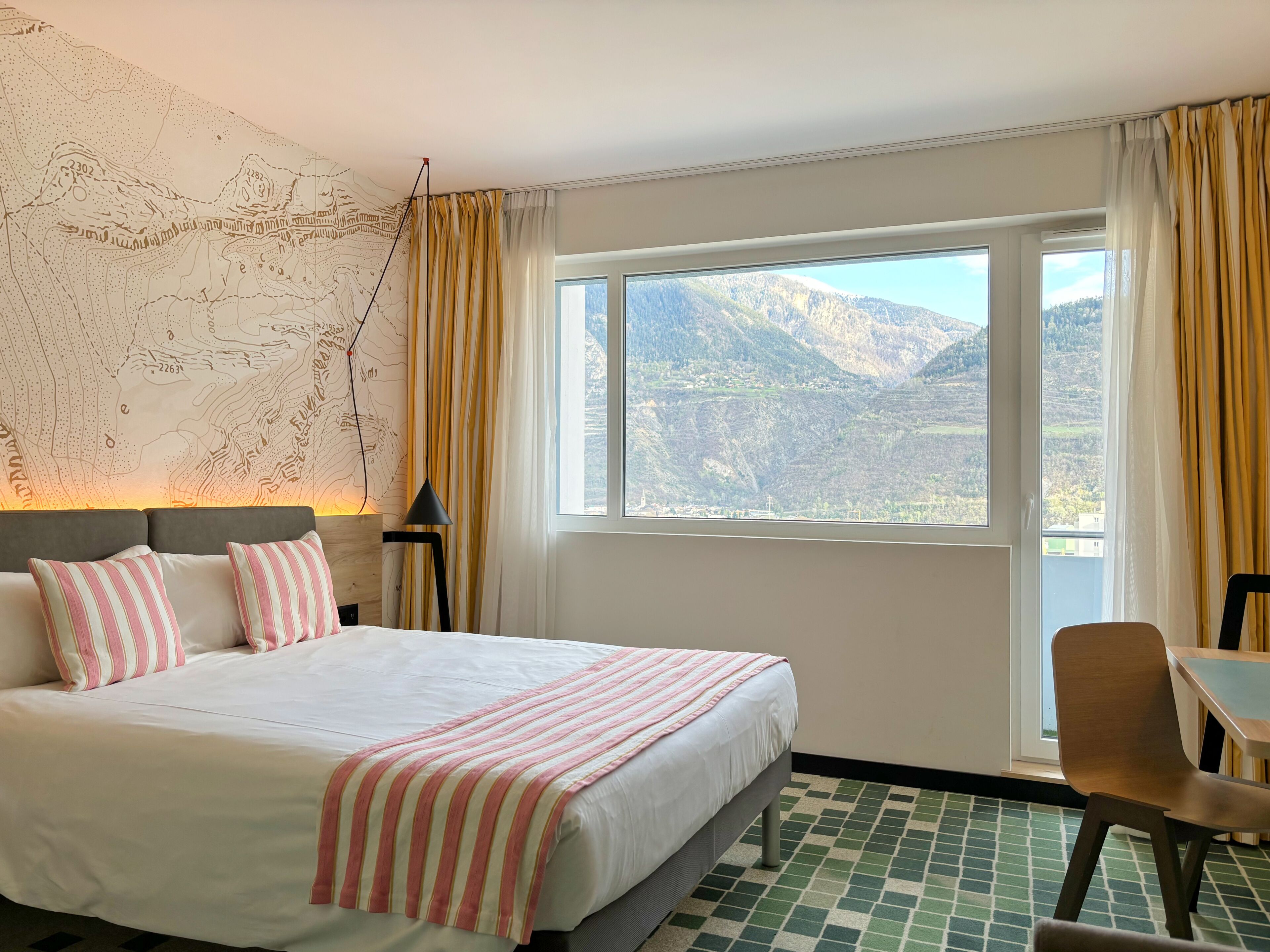 Een hotelkamer met een groot bed, een kaart aan de muur, uitzicht op bergen door het raam en een stoel bij het raam.